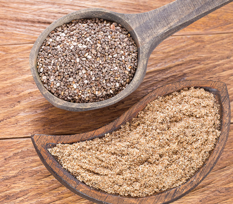 Whole chia seeds in a wooden spoon and ground chia in a bowl on a wood surface.